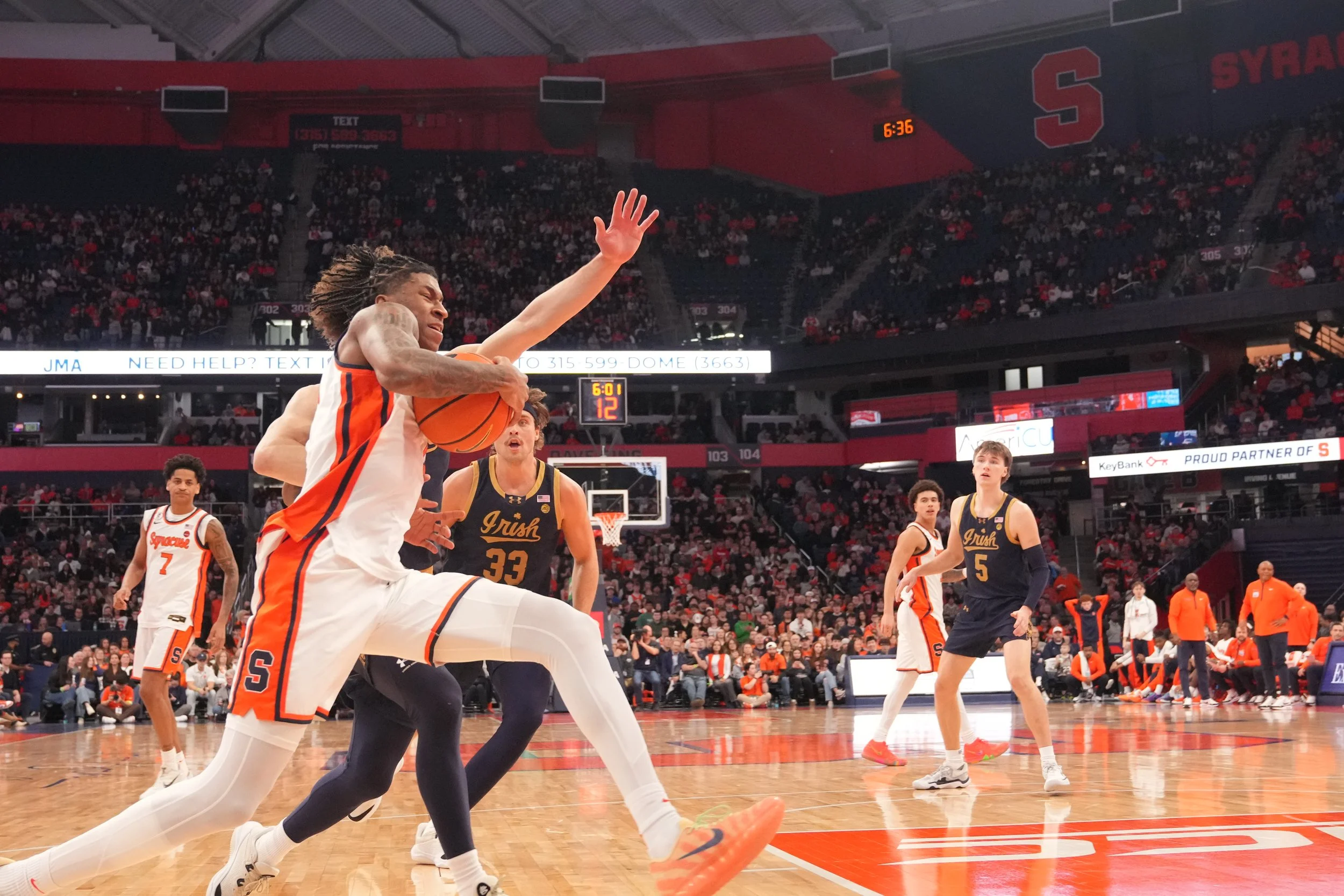 A basketball player in an orange and white uniform jumps to make a shot while a defender in a navy blue jersey attempts to block the shot in an indoor basketball arena filled with spectators.