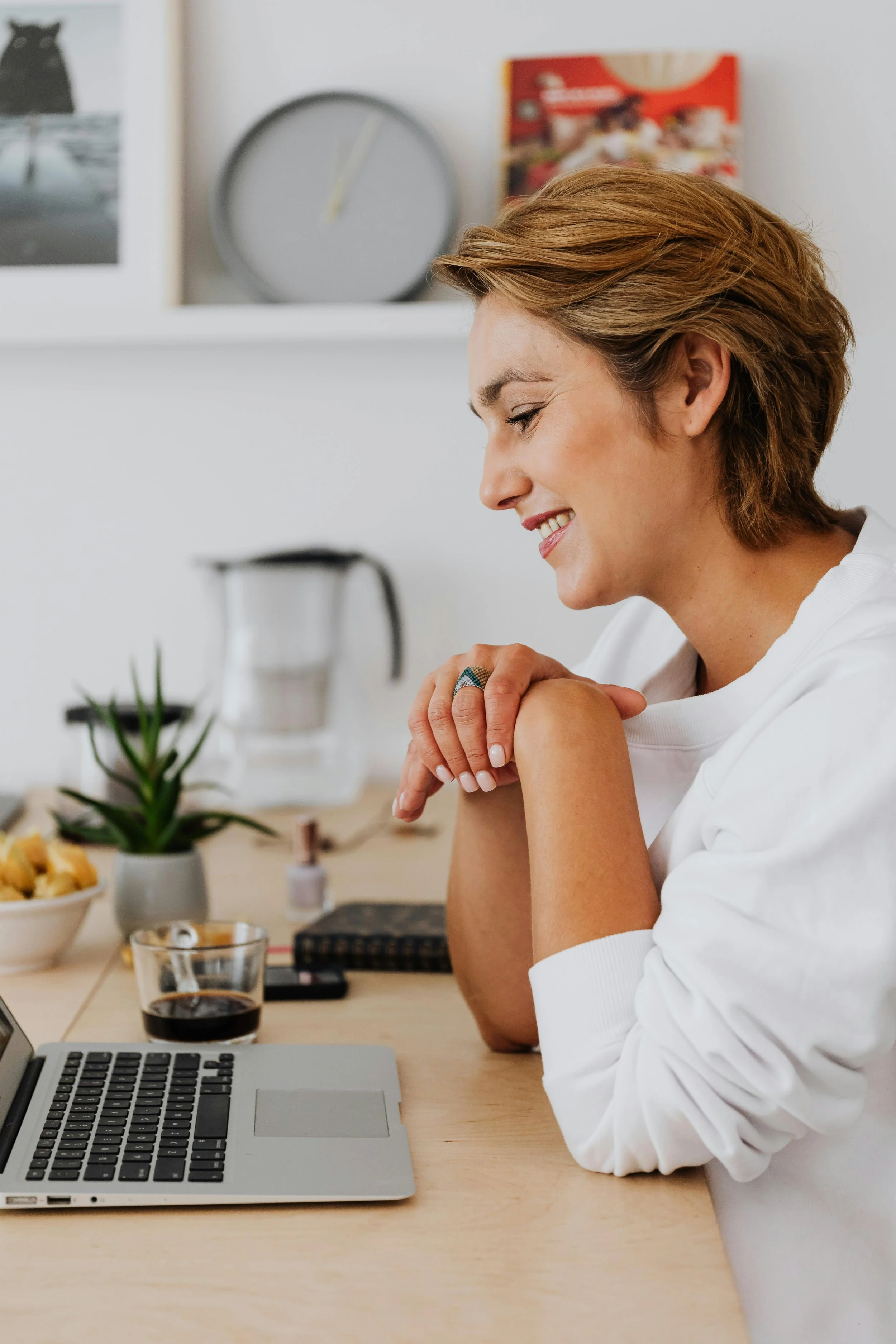 A woman smiling and sitting at a desk with a laptop, a glass of dark beverage, a potted plant, and various small items, in a bright room.