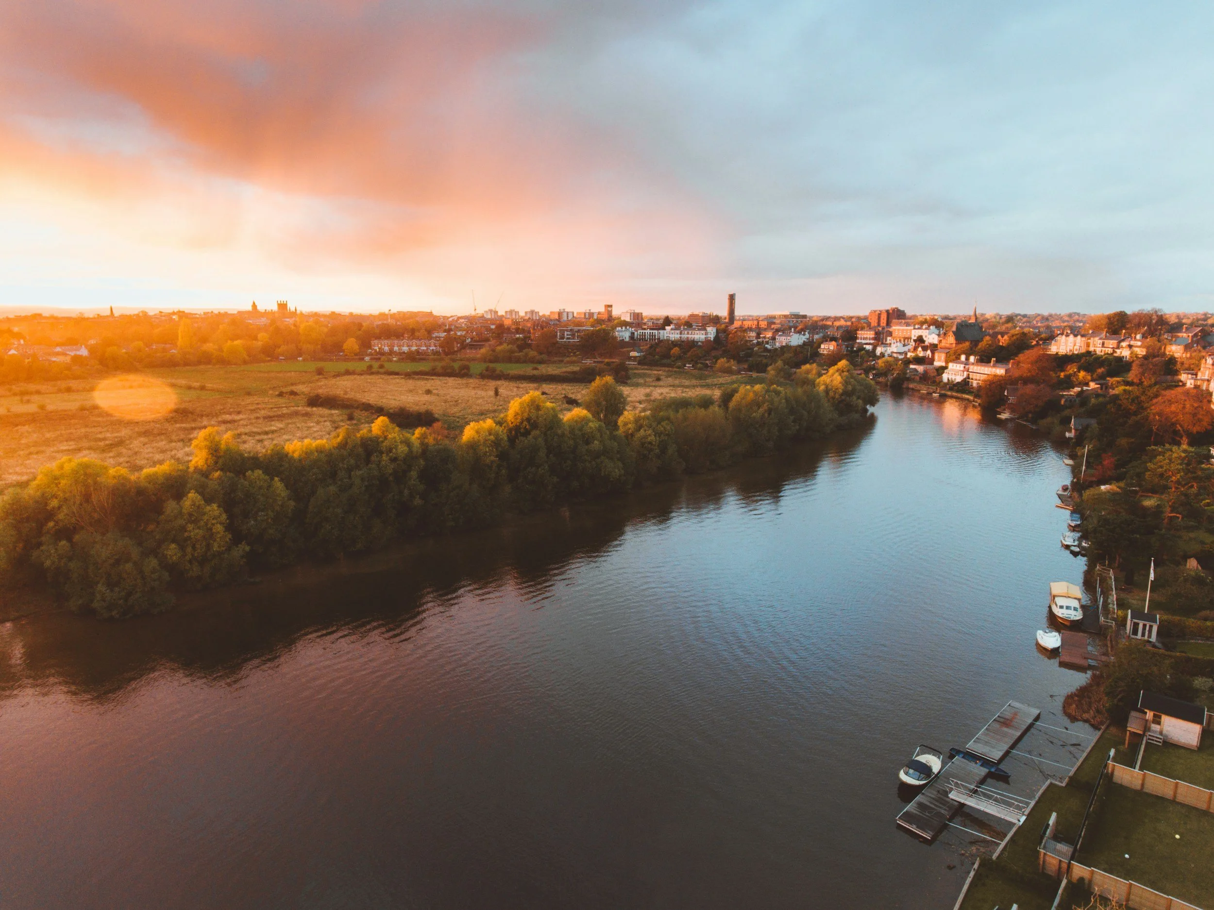 Sunset over a river with boats docked along the side, trees on both banks, and a city skyline in the distance.