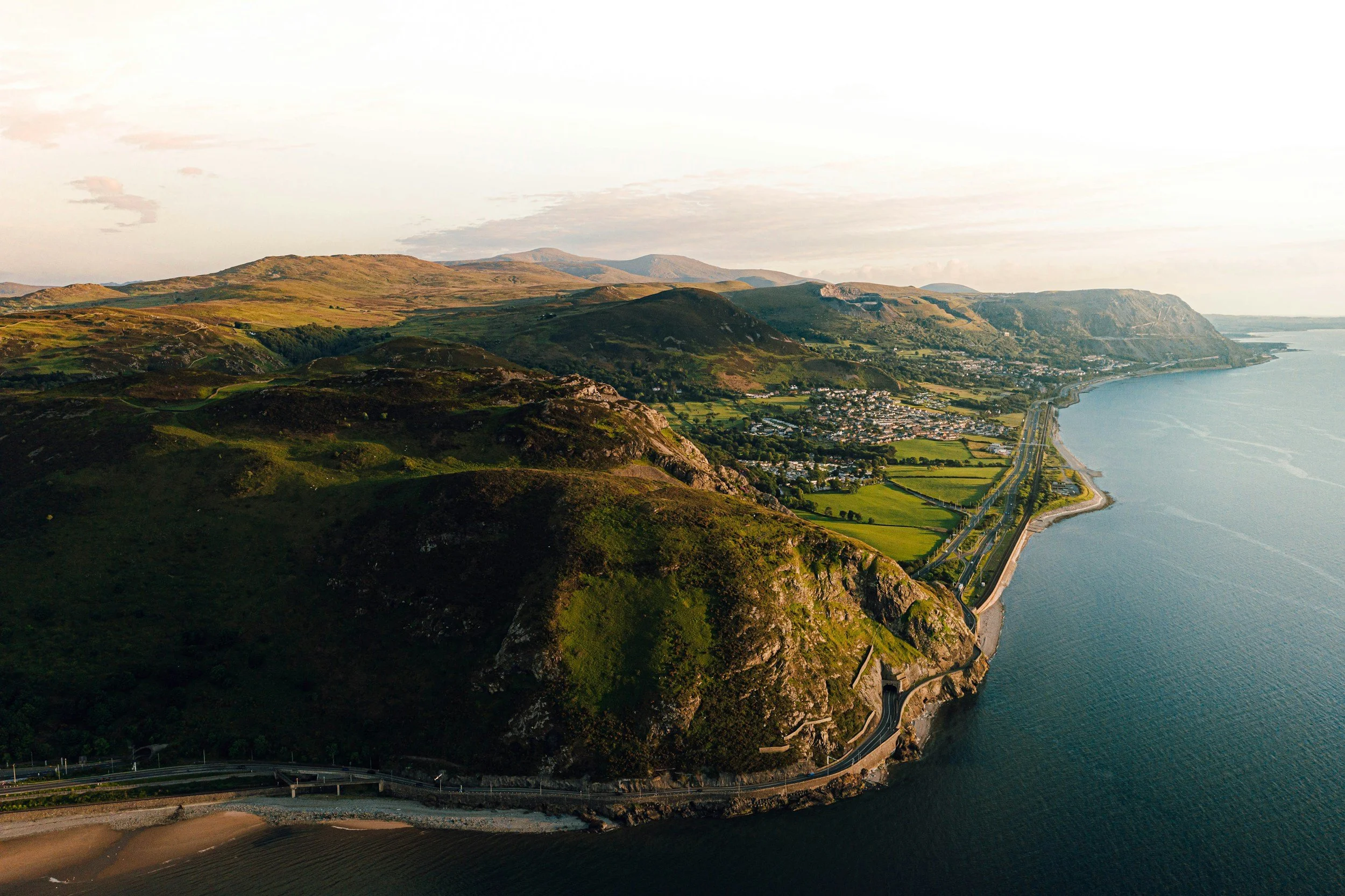 Aerial view of green hills and a coastline with a road running along the water, overlooking a town in the valley.