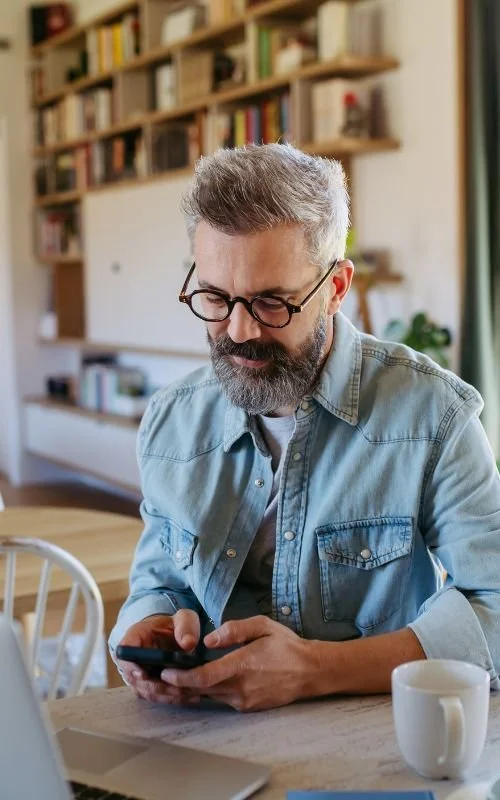 A middle-aged man with glasses and a beard looks at his phone, sitting at a wooden table in a cozy room with bookshelves in the background.