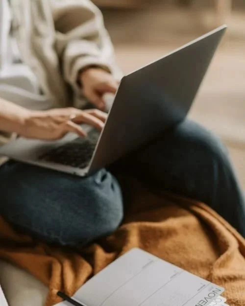 Person using a laptop on their lap with a notebook nearby.