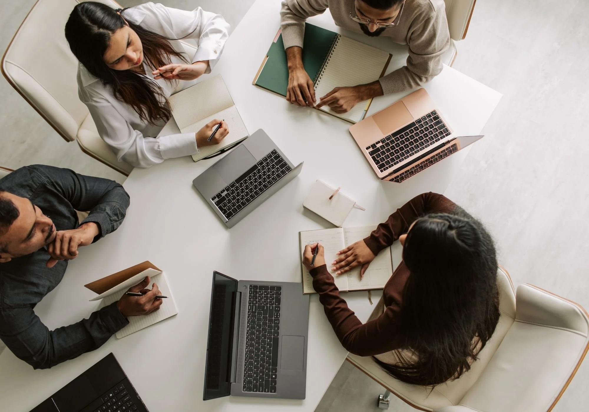 Top-view of four people sitting around a white table in a meeting, with laptops, notebooks, and pens. They are engaged in a discussion or brainstorming session.
