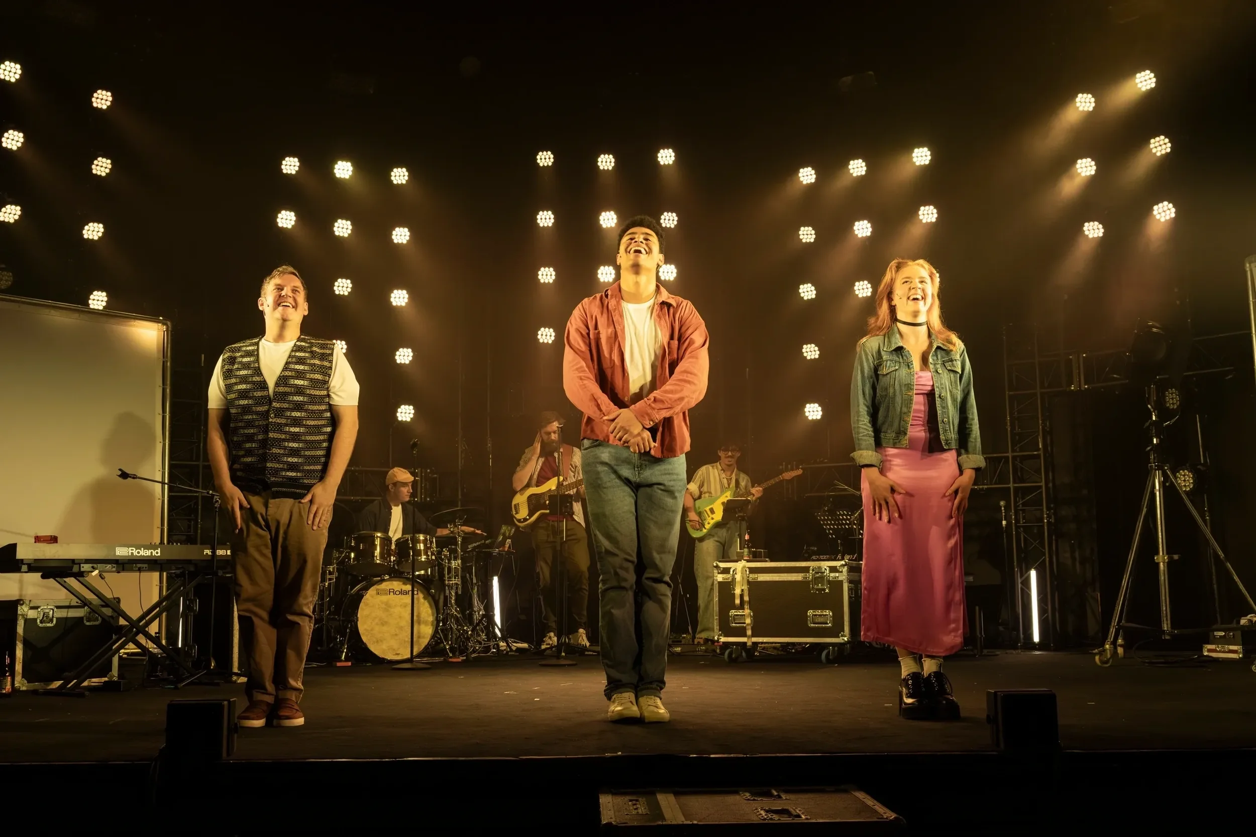 People standing on stage during a concert with bright stage lights in the background and a band playing instruments behind them.