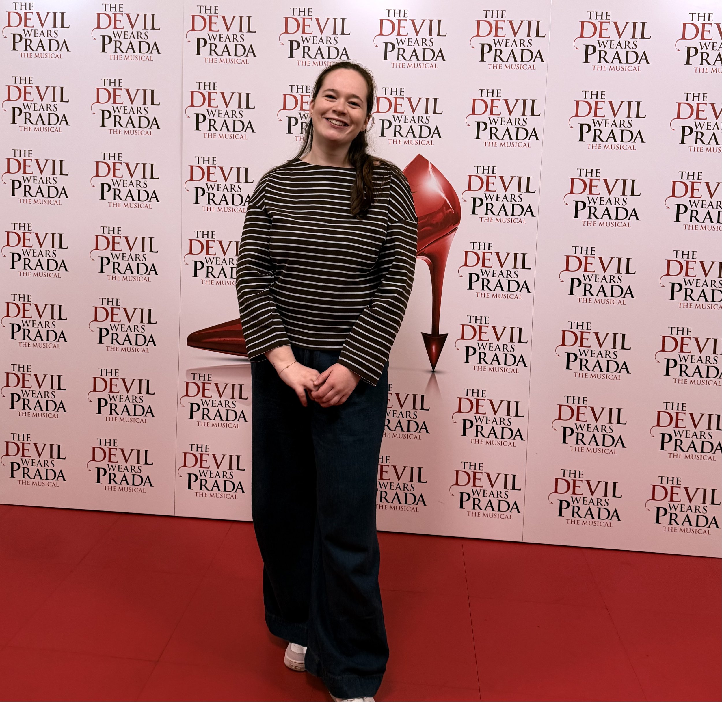A woman smiling at a promotional backdrop for 'The Devil Wears Prada: The Musical,' featuring a red high heel shoe design.