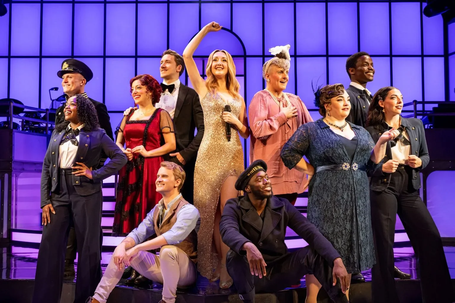 Cast of performers on stage in colorful costumes, celebrating after a show, with a purple-lit backdrop.