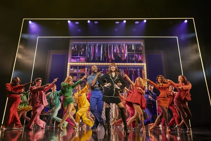 People performing on stage with a colorful wardrobe display in the background, colorful costumes, and stage lighting.