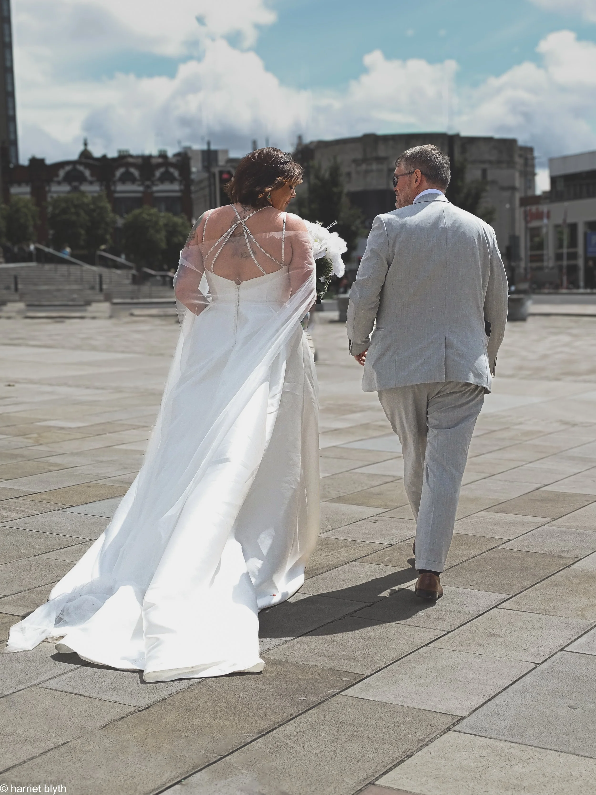 A bride and groom walking together outside on a paved area with modern buildings in the background, under a partly cloudy sky.