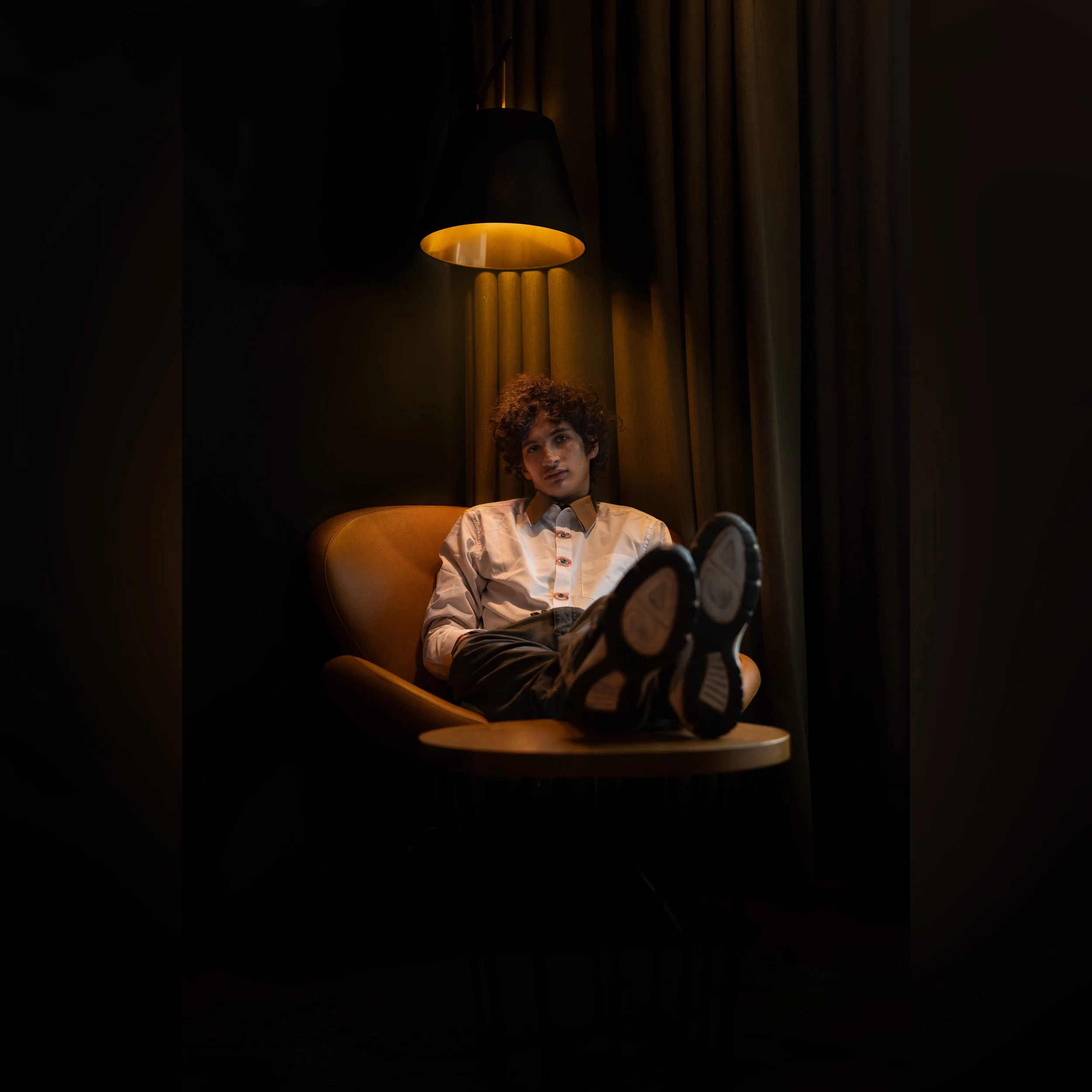 Person sitting on a yellow chair with feet up, illuminated by a warm yellow wall lamp, dark background with yellow curtain.
