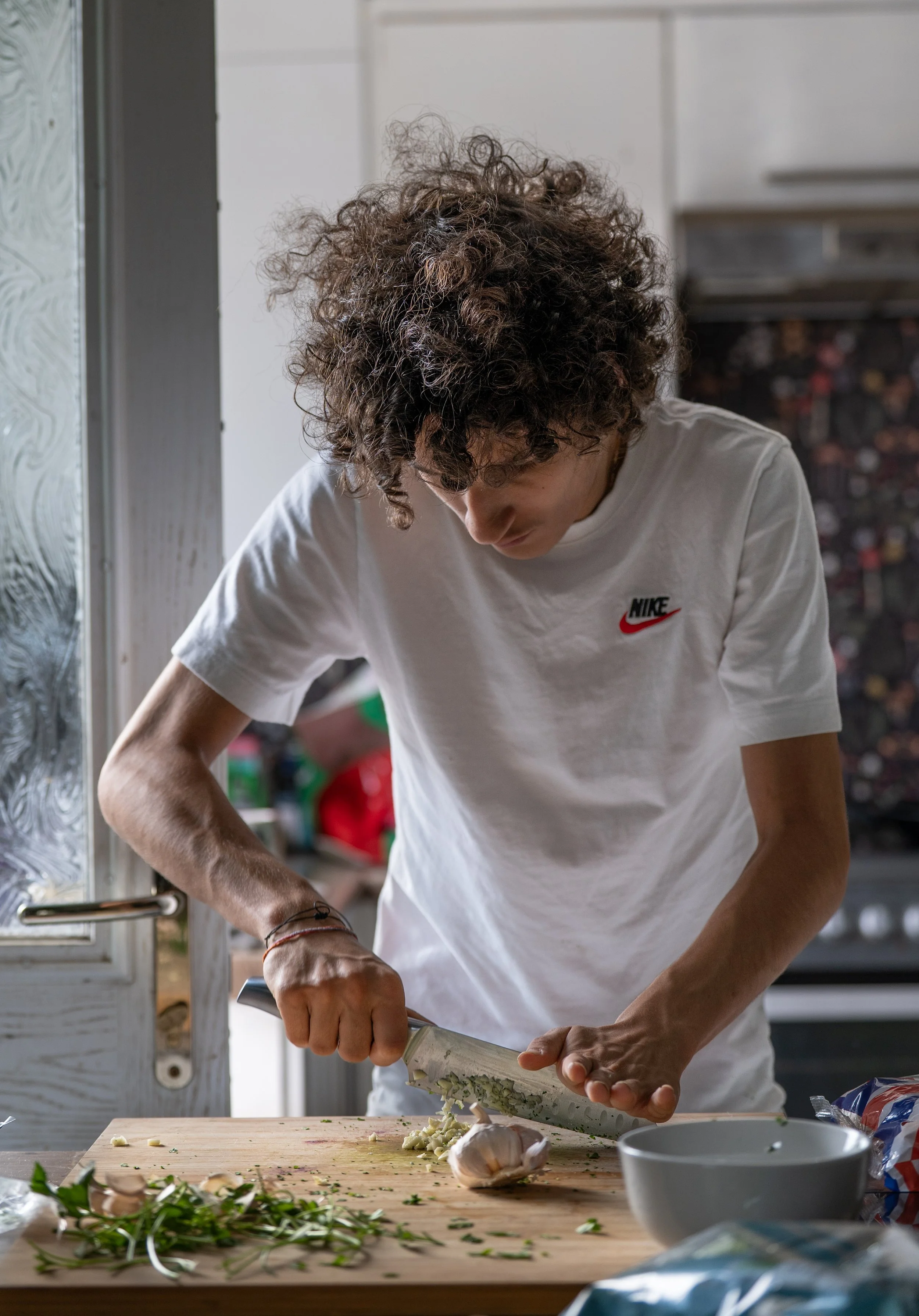 A person with curly hair wearing a Nike t-shirt is chopping vegetables on a wooden cutting board in a kitchen.