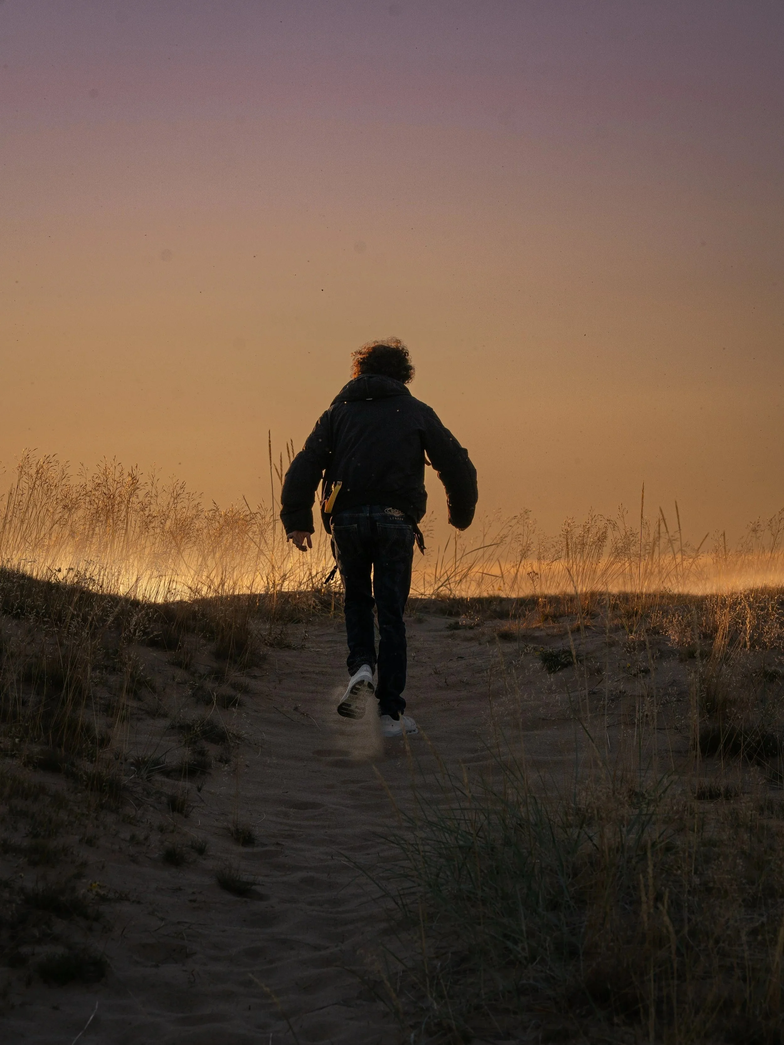 A person with curly hair wearing a dark jacket and jeans walking away on a sandy trail through tall grass during sunset or sunrise with a colorful sky.