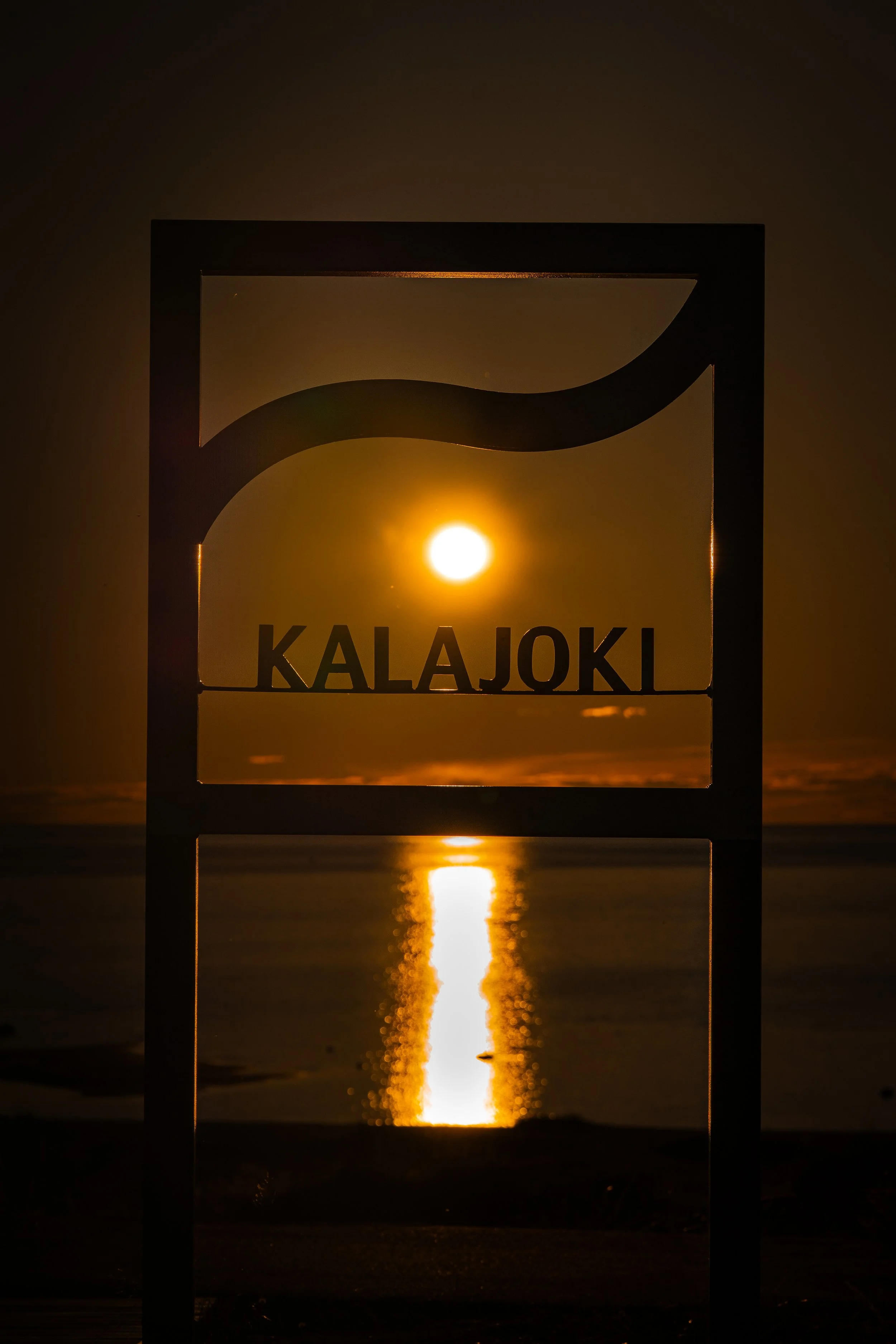 Sunset over the ocean viewed through a sign with the text 'KALAJOKI'. The sun's reflection creates a bright path on the water.