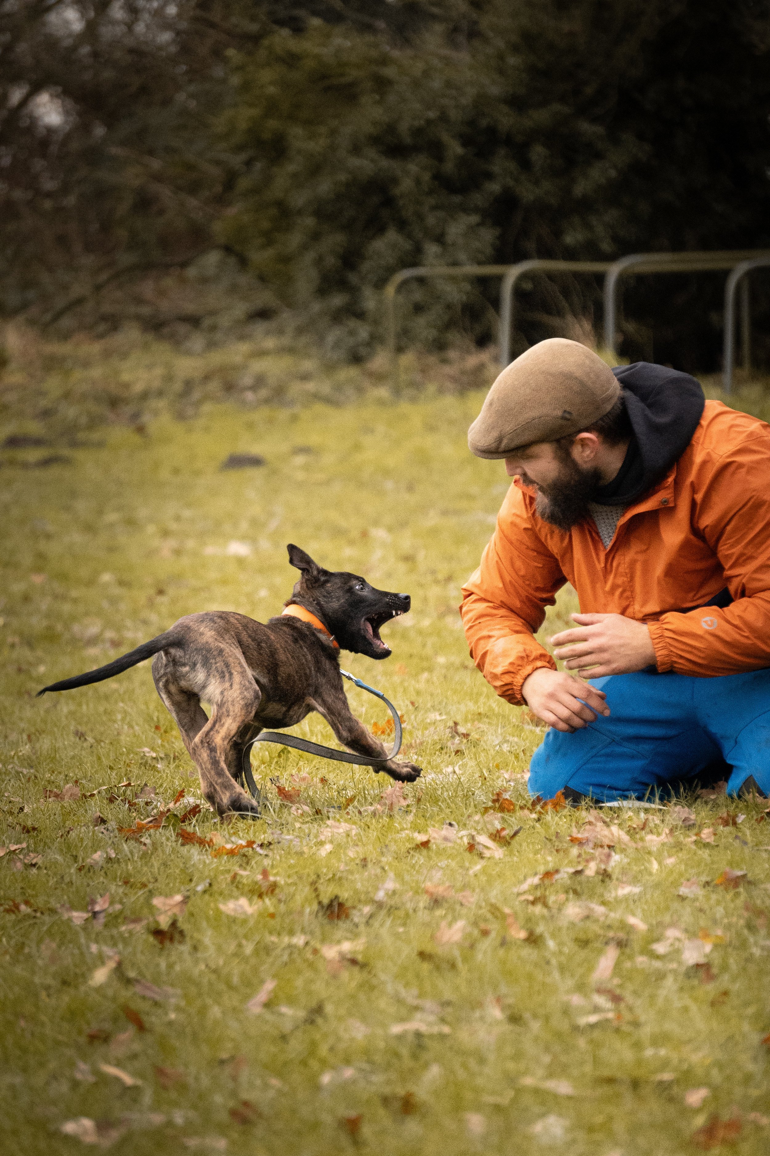 A man wearing a brown hat and orange jacket interacts with a black and brown dog on a leash in an outdoor park setting.