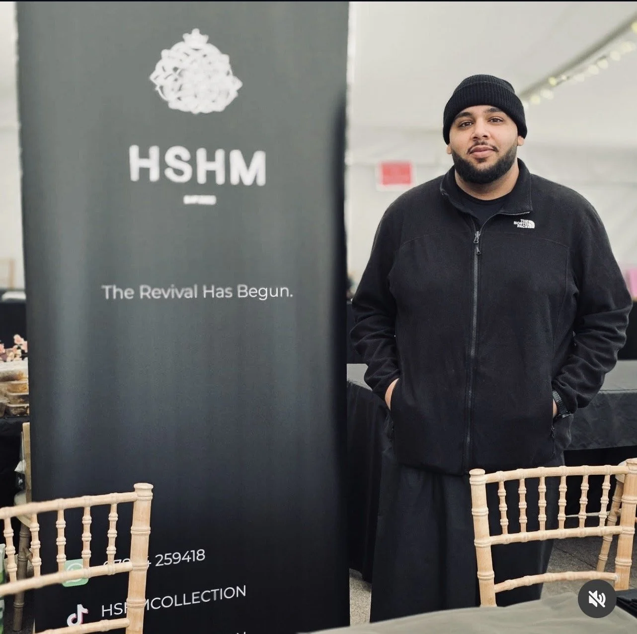 A man in a black North Face jacket and black beanie standing next to a black banner with white text that reads 'HSHM' and 'The Revival Has Begun.' The man has a beard and is looking at the camera, with hands in his pockets.