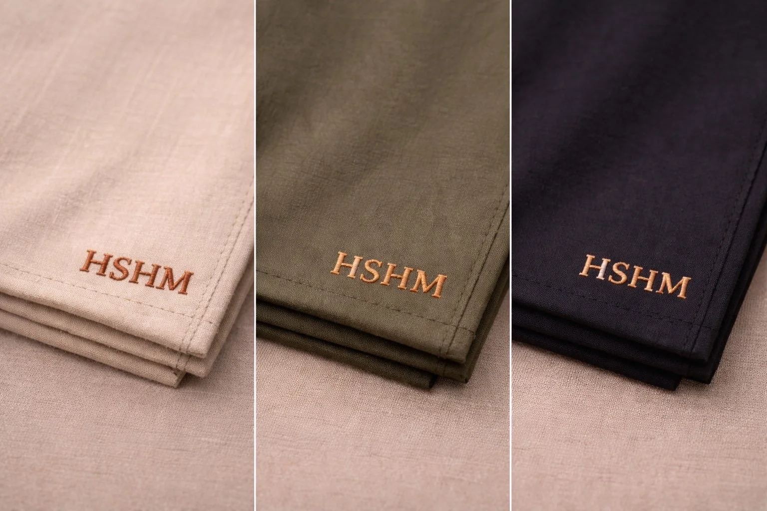 Set of three folded fabric napkins in beige, olive green, and black with embroidered HSHM logo in copper thread on each napkin.