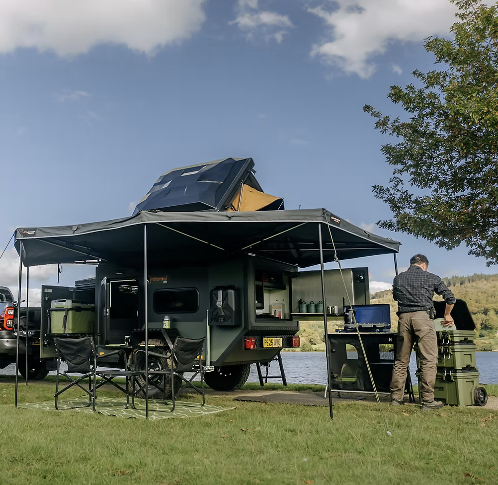 A man cooking on a barbecue grill next to a camper trailer by a lake, with outdoor furniture and a tree.