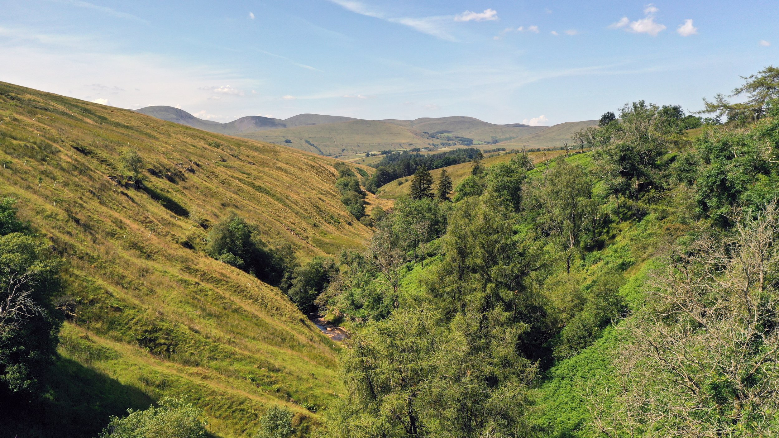A scenic view of rolling hills with green vegetation, a small river flowing through a valley, and a clear blue sky with a few clouds.