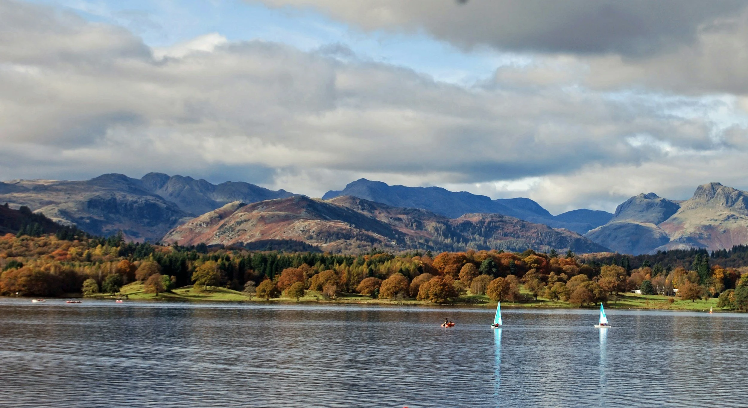 A lake with small sailboats, surrounded by autumn-colored trees and mountains in the background under a partly cloudy sky.