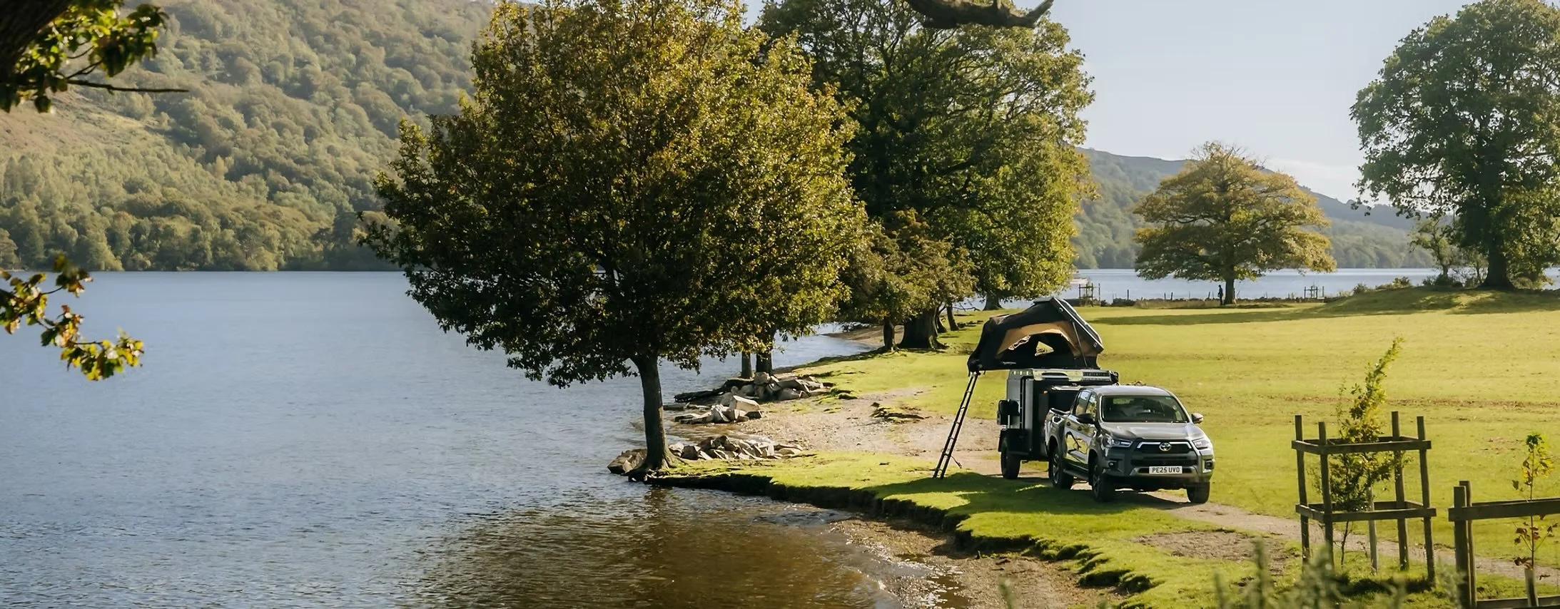 A lakeside scene with green trees lining the water, a grassy area with a parked truck and rooftop tent, and mountains in the background.
