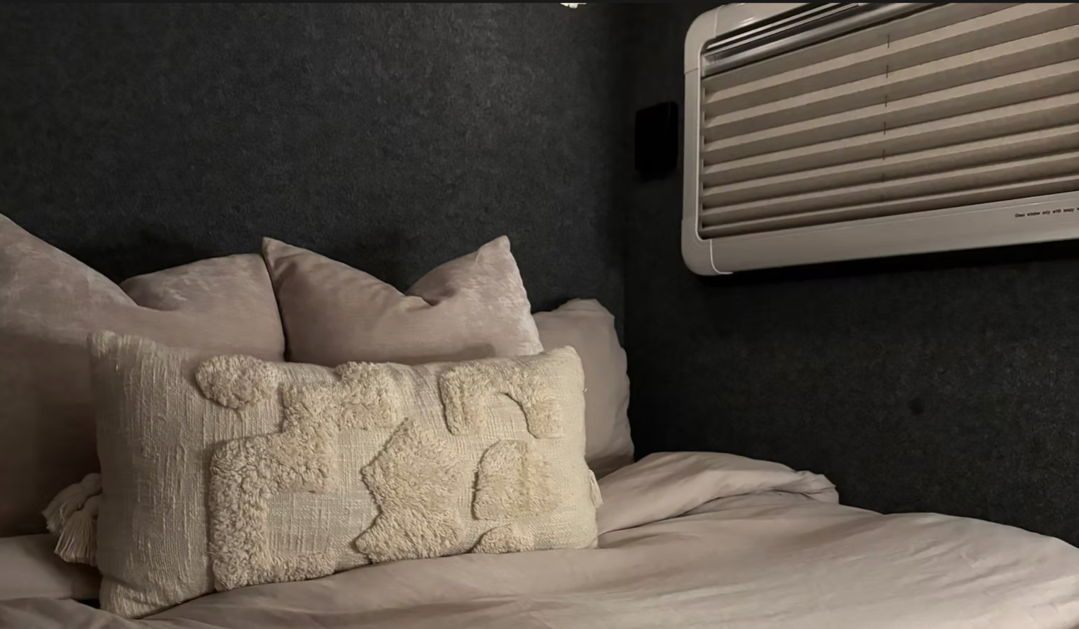 A bed with multiple pillows against a dark wall, with an air conditioning unit mounted on the wall above the bed.