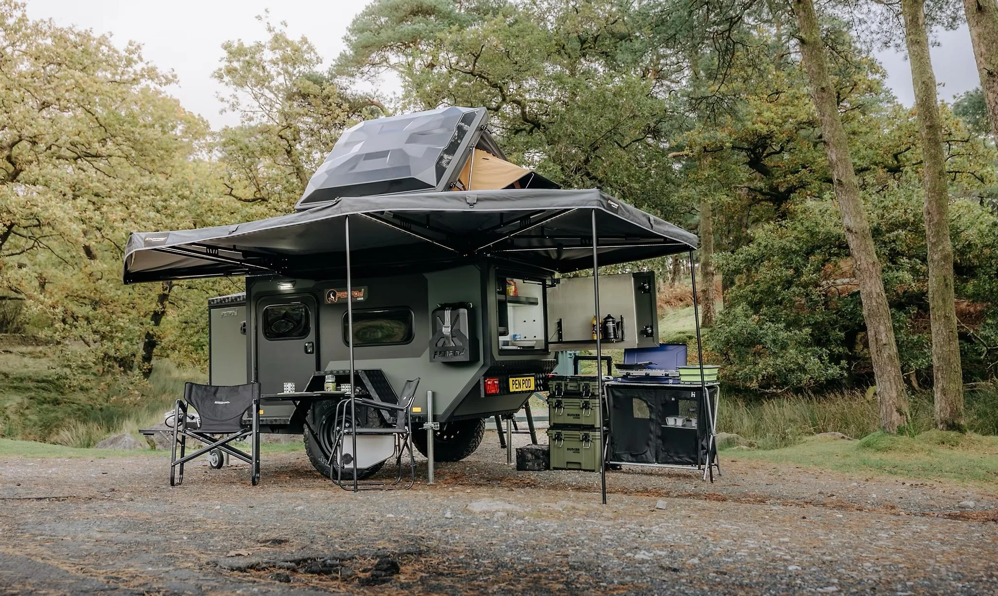 A travel trailer converted into a camper with an outdoor canopy set up in a wooded area during daytime.