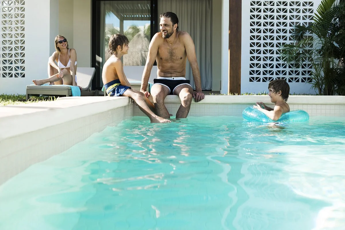 A family of four enjoying time by a swimming pool. The father and son are sitting on the pool's edge with their feet in the water, the father smiling at the son. The mother and daughter sit on lounge chairs nearby, smiling and relaxing, on a sunny day.