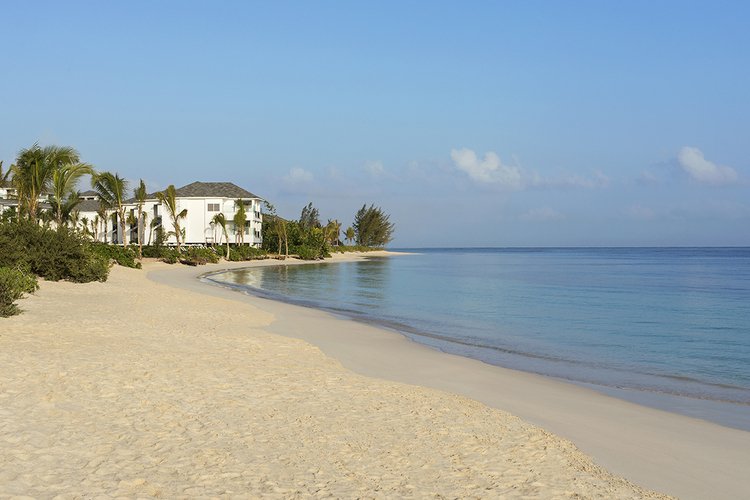 Sunset view of a peaceful beach with white sand, calm ocean water, and a few resort buildings with palm trees.