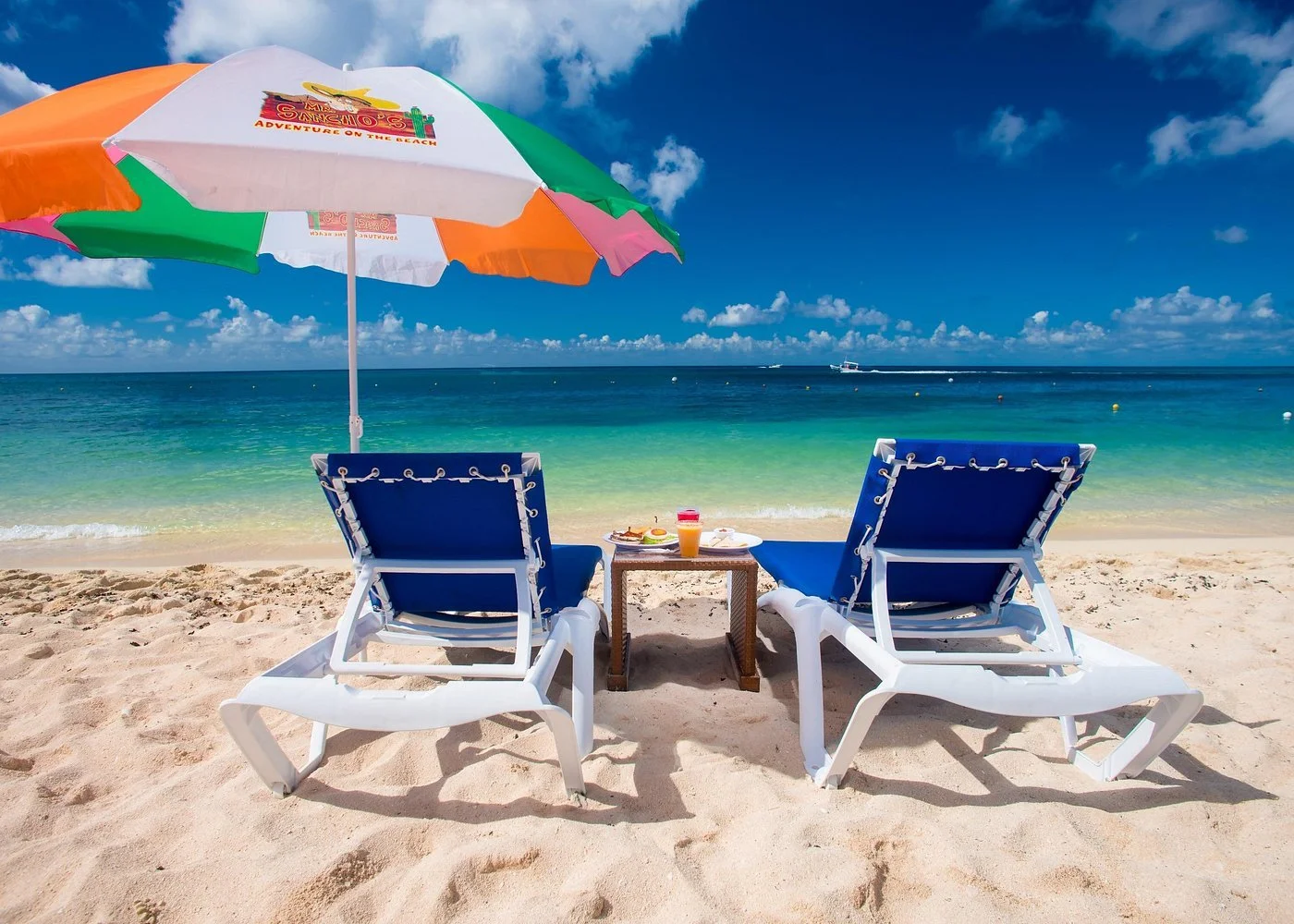 Beach scene with two blue lounge chairs, a small table with breakfast items and a colorful umbrella, overlooking the ocean with a boat in the distance under a partly cloudy sky.