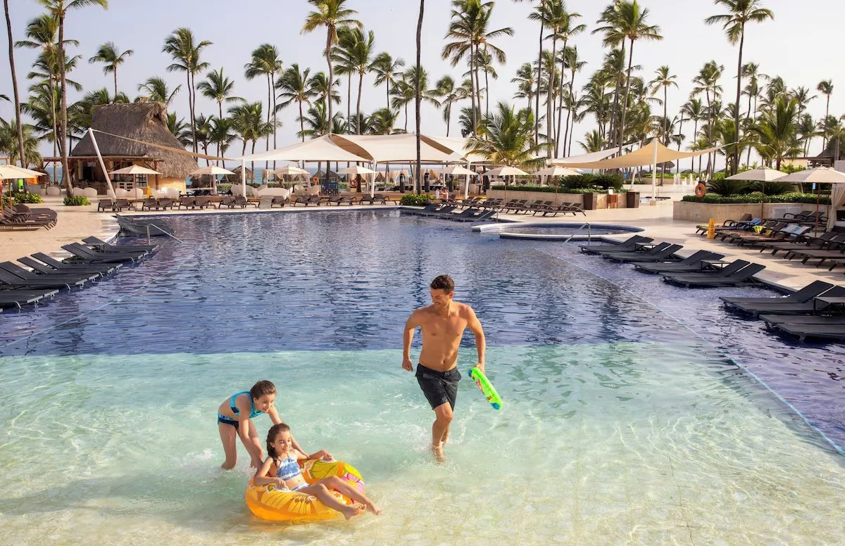A man and two children enjoying a swimming pool at a tropical resort with palm trees, lounge chairs, umbrellas, and thatched-roof structures in the background.