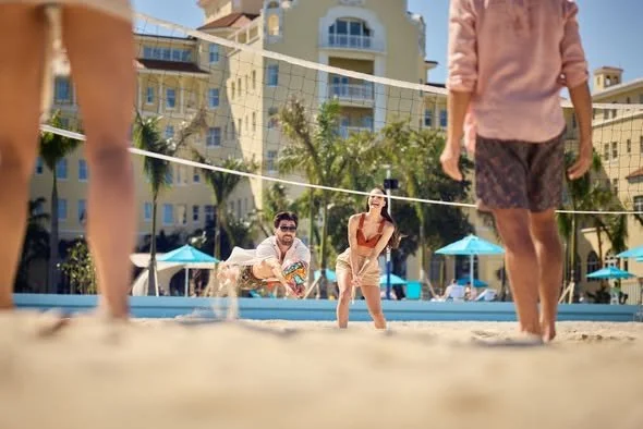 People relaxing and playing volleyball on the beach with hotel buildings and palm trees in the background.