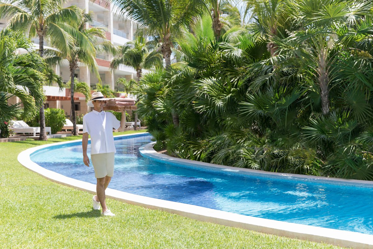 A woman in a white shirt, beige shorts, and a straw hat walking beside a curved swimming pool in a tropical resort. She is carrying folded towels on her shoulder and the surroundings include palm trees and lounge chairs.