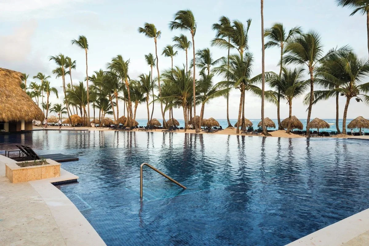Poolside view with palm trees, thatched umbrellas, and lounge chairs overlooking the ocean.