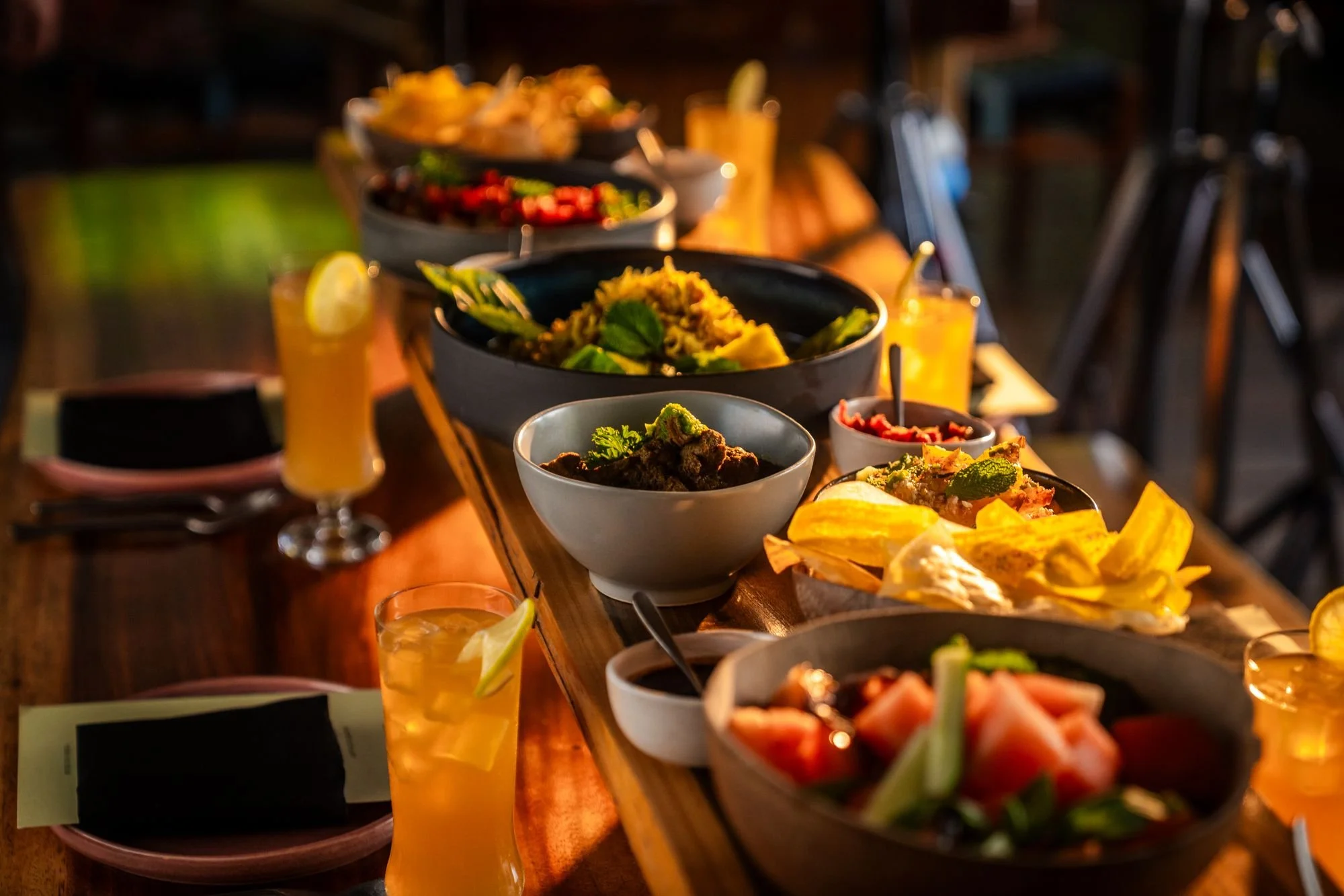 A long wooden table with bowls of various colorful foods and glasses of yellow drinks garnished with lemon slices, set for a meal in warm lighting.