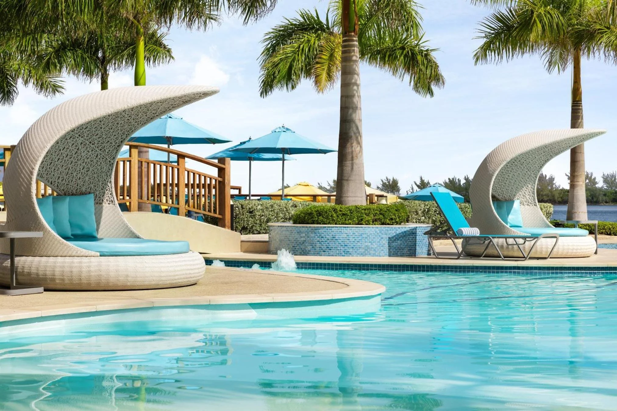 Luxury swimming pool with curved cabana chairs, tropical palm trees, and umbrellas in a resort setting.