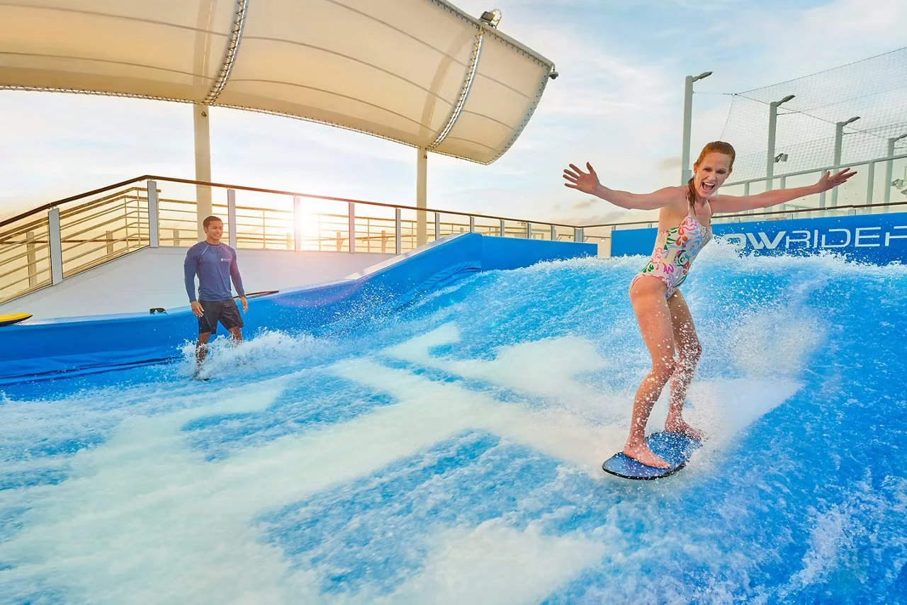 A woman in a colorful swimsuit surfing on an indoor wave pool with a man in the background.
