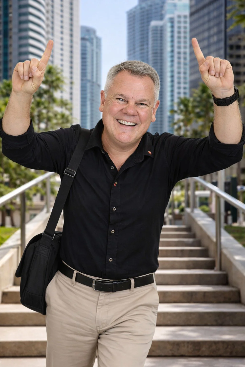 A smiling middle-aged man with gray hair, wearing a black shirt and beige pants, standing on outdoor stairs in an urban setting, pointing upwards with both hands.