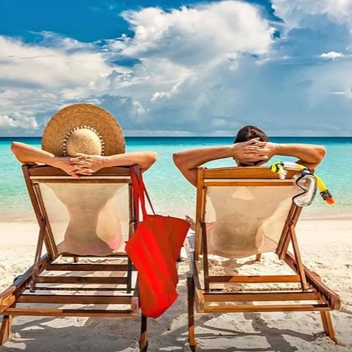 Two people sitting on lounge chairs at the beach facing the ocean, one with a wide-brimmed hat and the other with sunglasses, under a partly cloudy sky