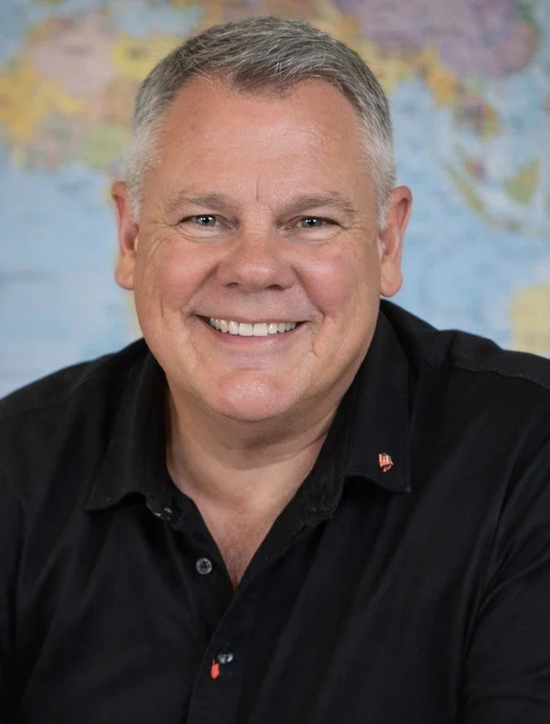 A smiling middle-aged man with short gray hair wearing a black shirt, sitting in front of a blurred world map background.