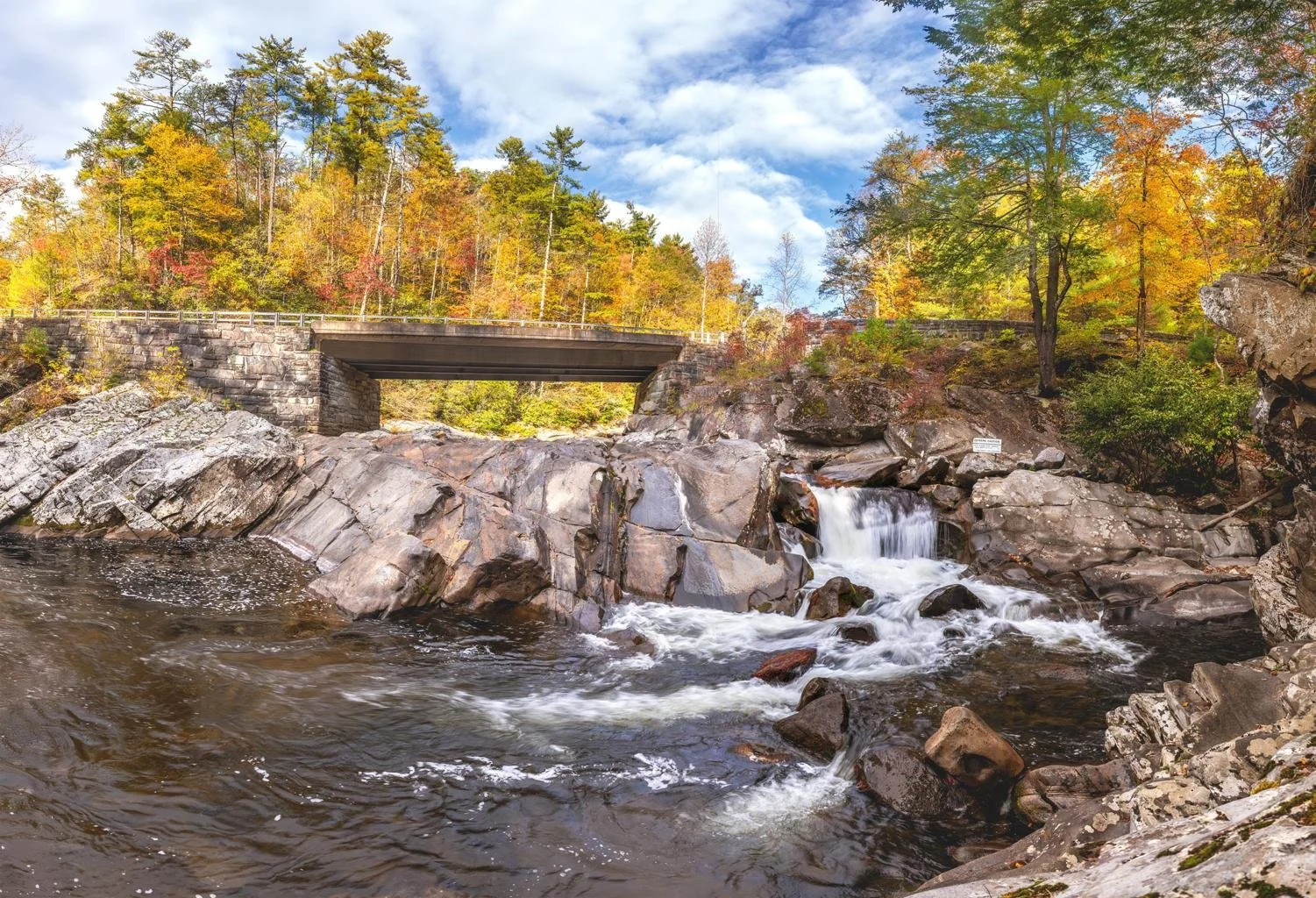 A river with a small waterfall flowing over rocks, with a bridge and colorful autumn trees in the background under a partly cloudy sky.