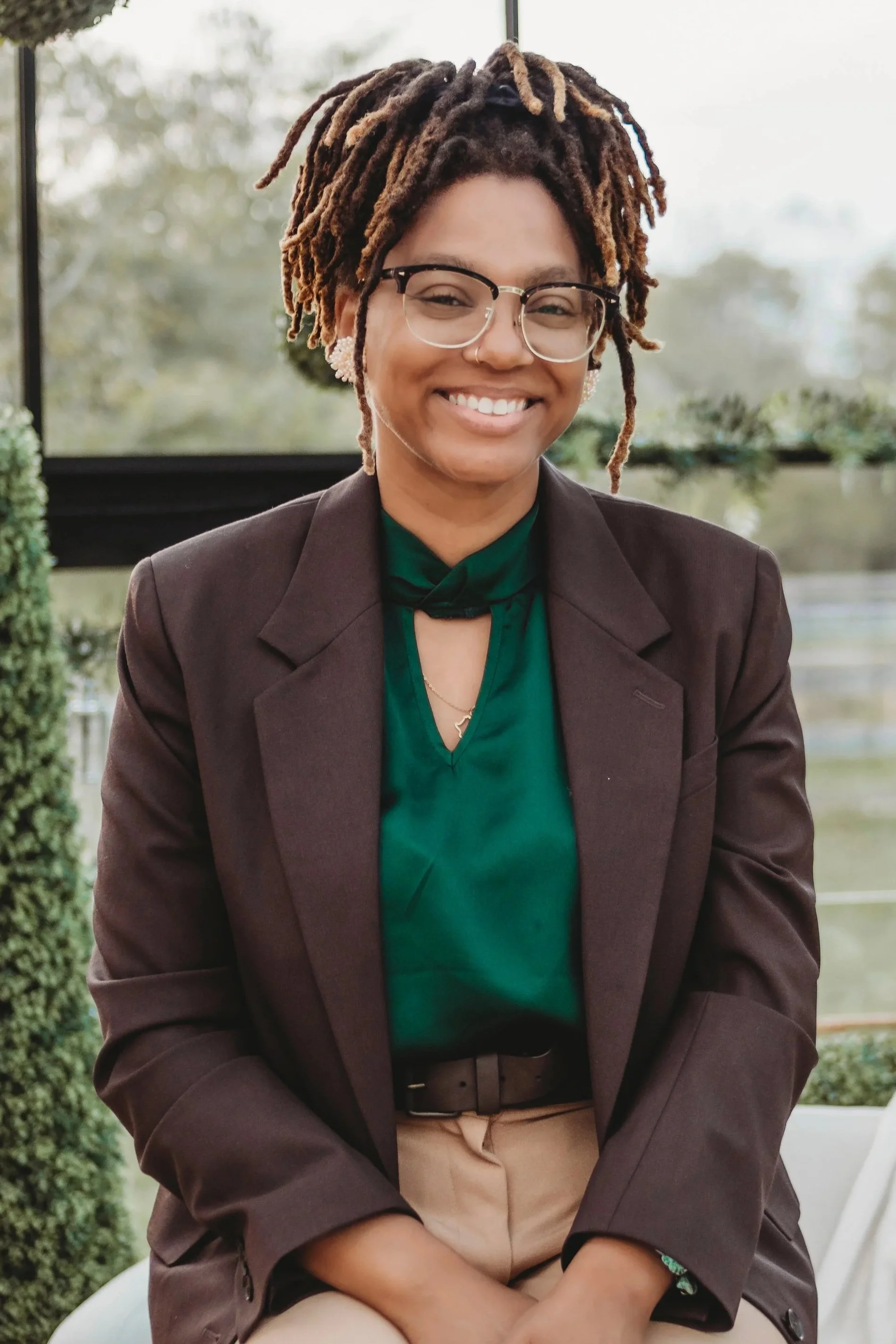 A woman with glasses and dreadlocks smiling, wearing a dark blazer over a green blouse, sitting outdoors by a window with blurred greenery in the background.