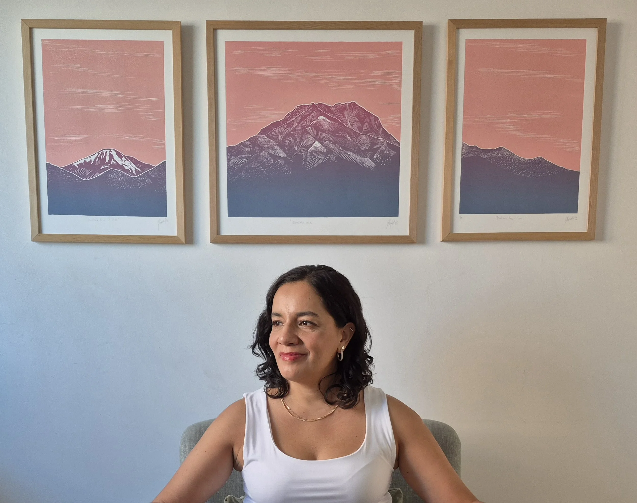 Mujer sentada en una silla frente a una pared con tres pinturas de montañas en fondos rosados.