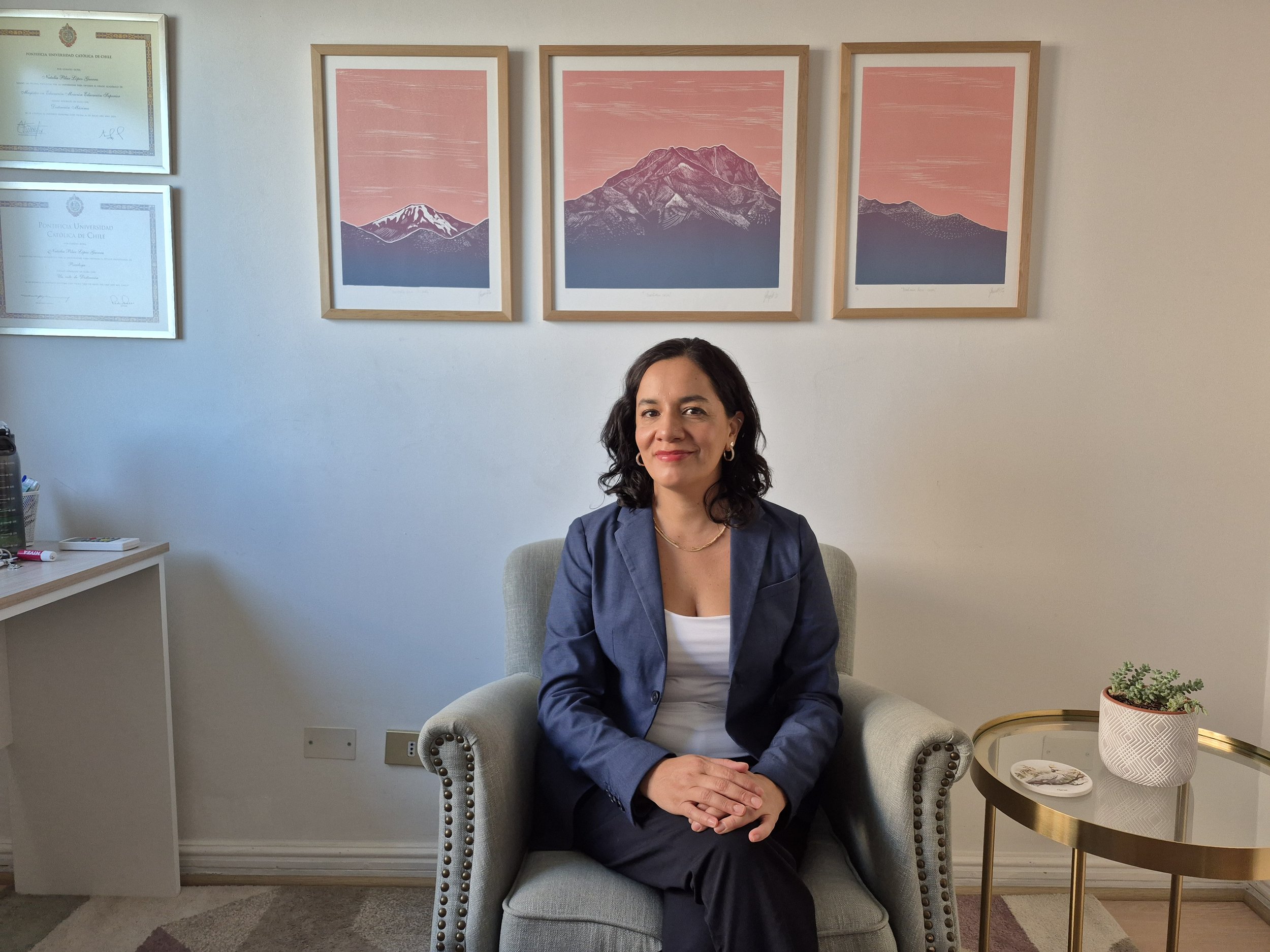 Una mujer sentada en una sillón gris en una oficina, con cuadros de montañas en la pared y una mesa dorada con una planta y una placa.