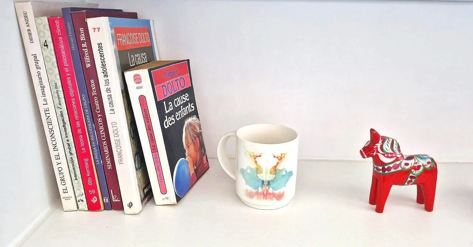 Estantería con libros, una taza blanca con diseño colorido, y una figura de caballo de madera decorativo en rojo.