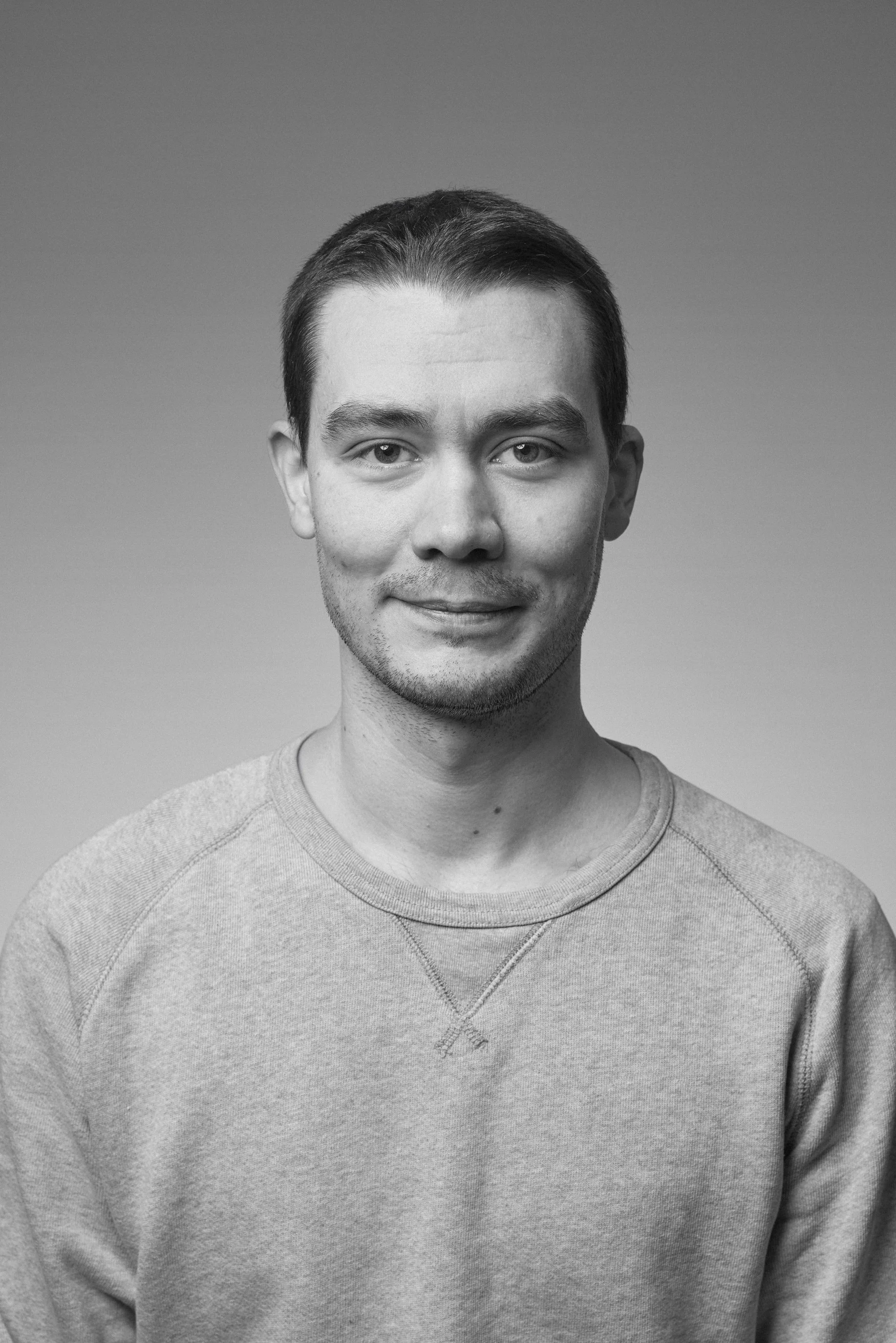 Black and white portrait of a young man with short dark hair, wearing a casual sweatshirt with a decorative seam across the chest, smiling softly against a plain gradient background.