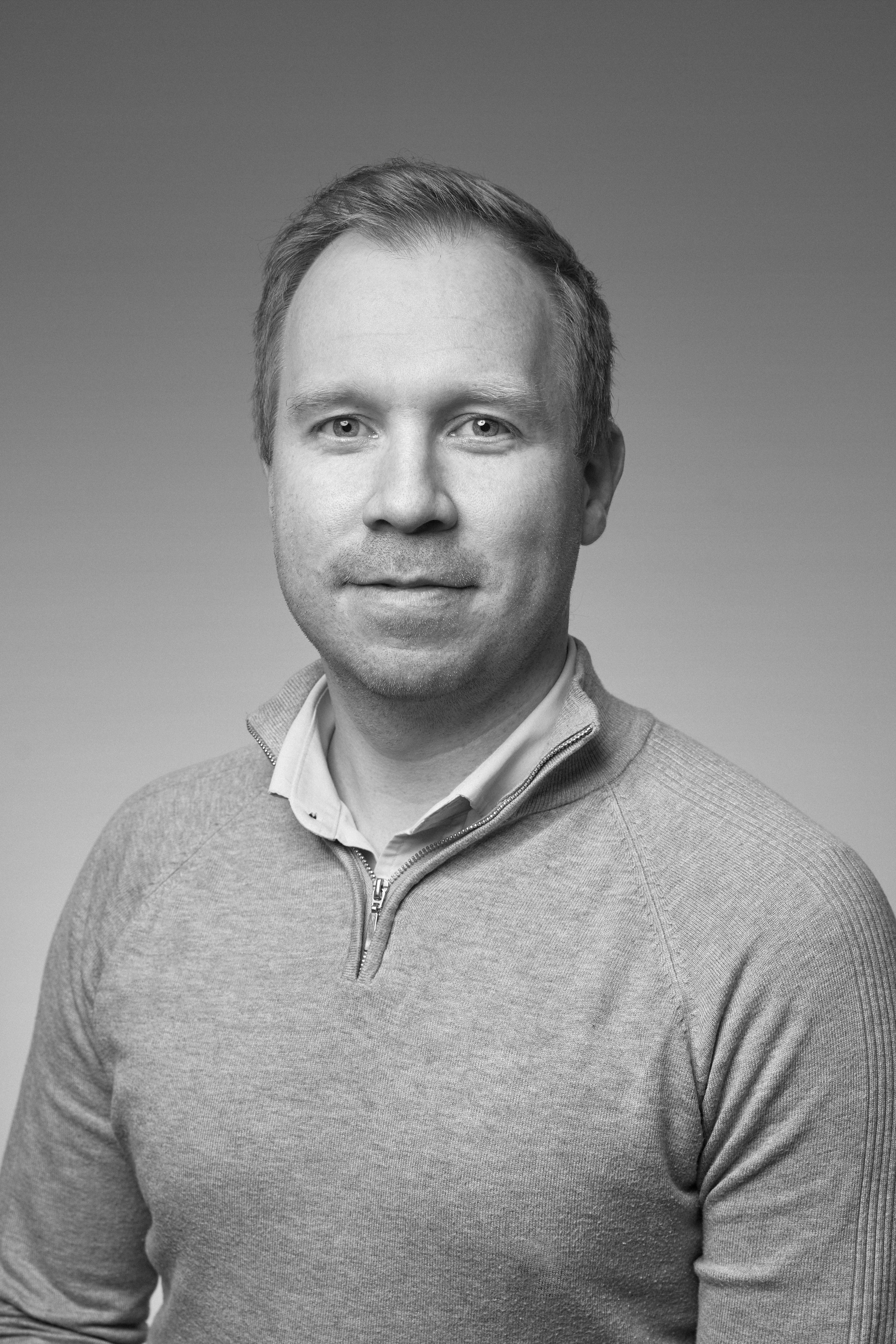 Black and white portrait of a man with short hair, wearing a zip-up sweater over a collared shirt.