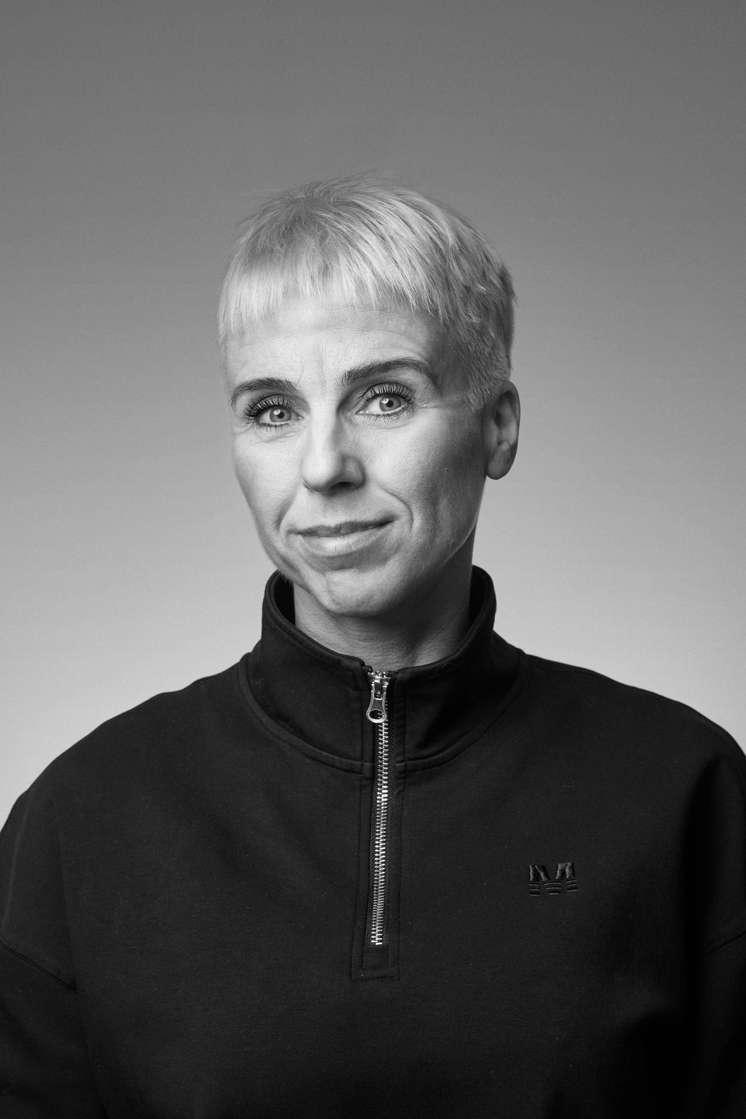 Black and white portrait of a woman with short blonde hair, wearing a black zip-up jacket with a small logo on the chest, against a plain background.