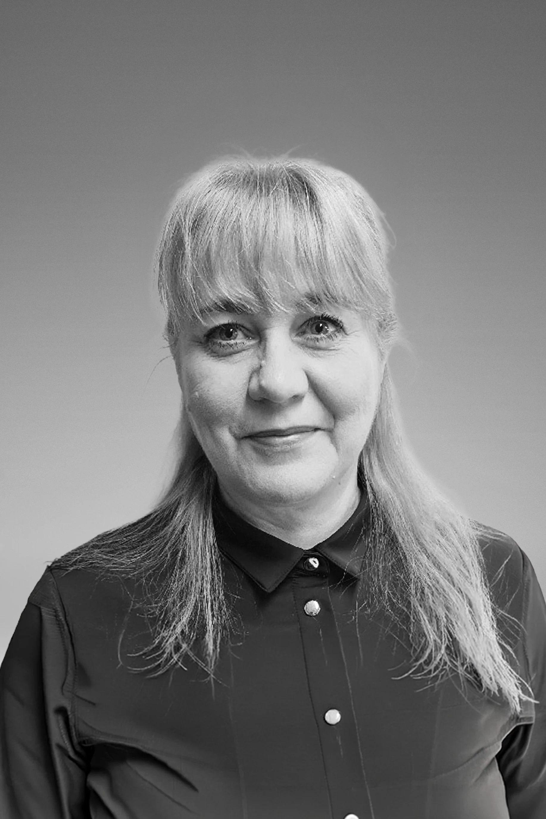 A woman with long, light-colored hair styled with bangs, smiling, wearing a dark collared shirt with metallic buttons, in black and white.