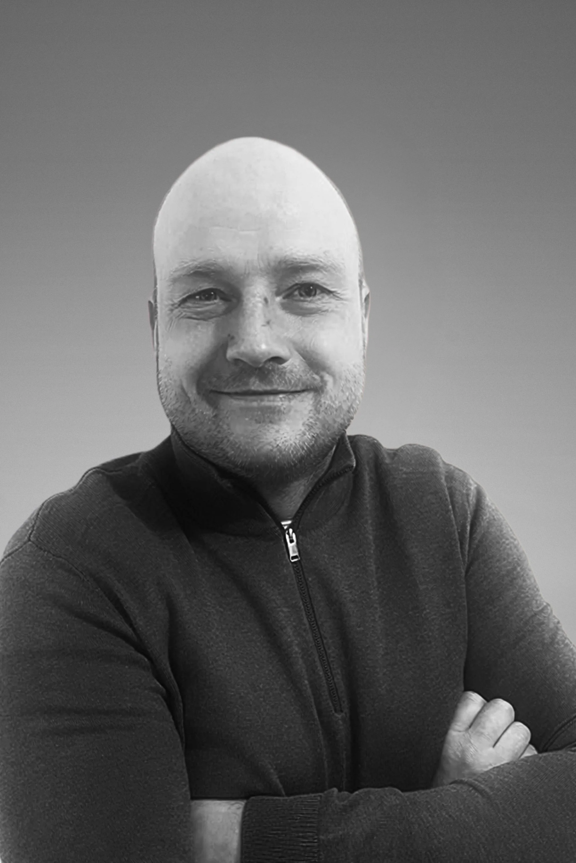 A black-and-white photo of a man with a shaved head and a slight smile, wearing a zip-up jacket, with his arms crossed on a table and the background fading to a plain gradient.
