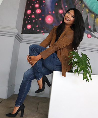 A woman sitting on a white platform, smiling and posing in front of a colorful, abstract background. She is wearing a brown jacket, blue jeans, and black heels.