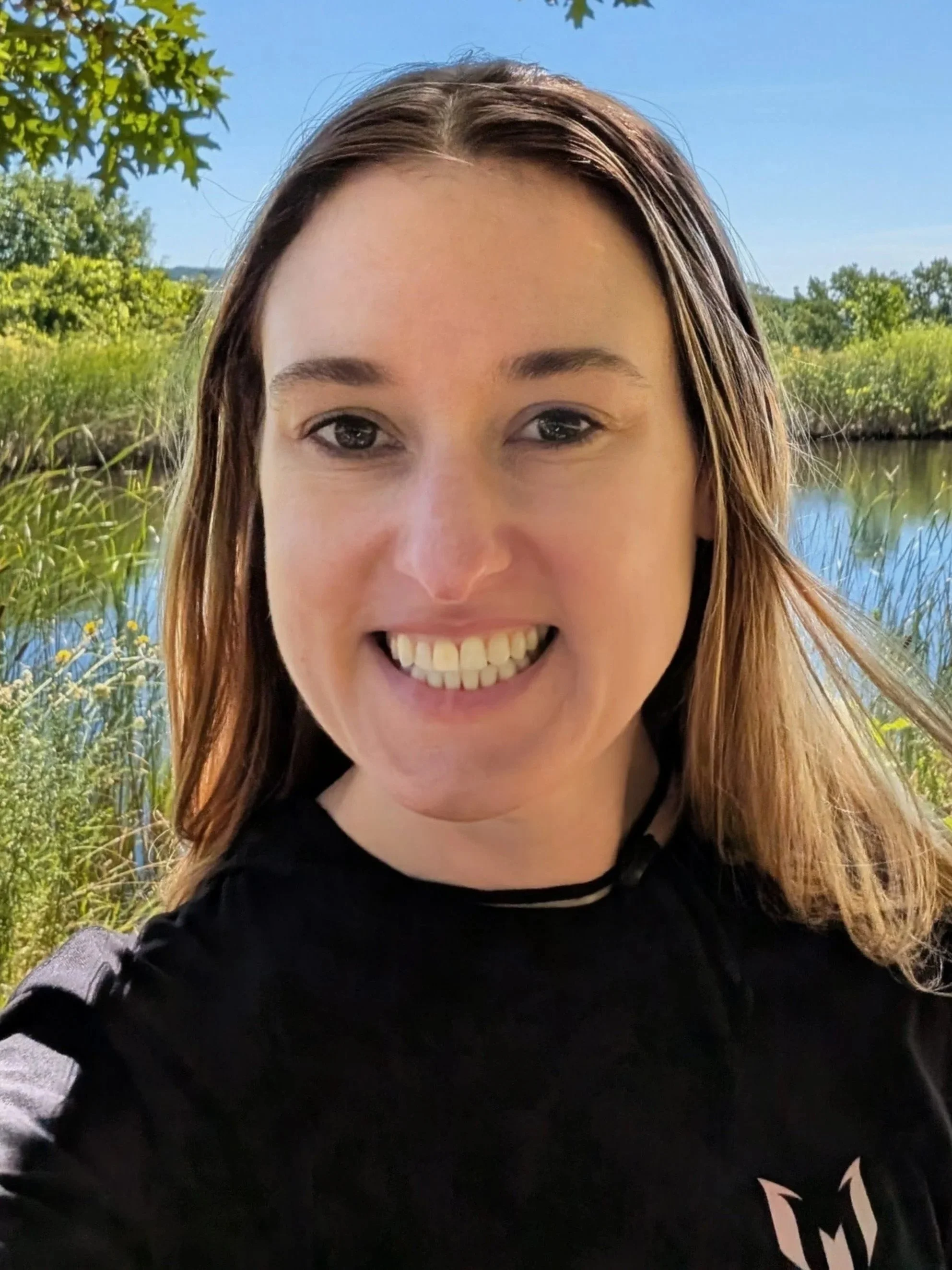Smiling woman in black shirt outdoors near a body of water with green foliage and trees in the background.